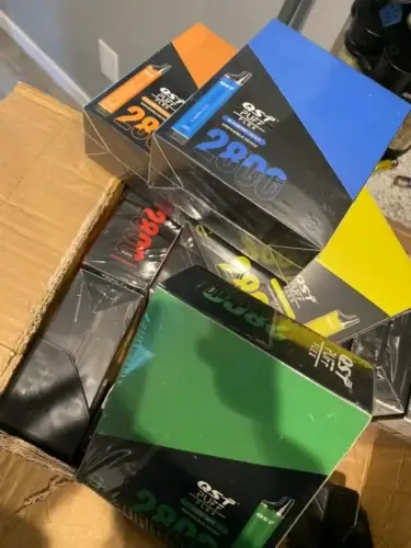 A cardboard box containing multiple packages of disposable vape devices marked with the brand name "Puff Flex 2800" in various colors such as orange, blue, green, and yellow.