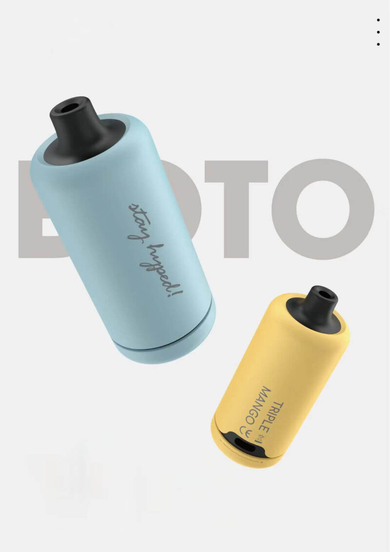 Två färgglada vape-pennor, en ljusblå med "stay hyped!" text och den andra gula märkt "TRIPLE MANGO", placerad mot en vanlig bakgrund med suddig text "BOTO."