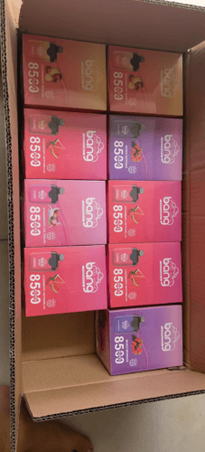 A cardboard box containing multiple boxes of Bang disposable vape devices in various flavors including Strawberry Banana, Mixed Berries, Strawberry Ice Cream, and Strawberry Watermelon, each labeled with "8500 PUFFS."