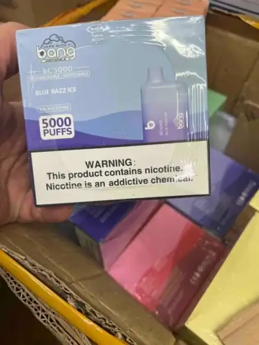 Una mano sosteniendo una caja de vapeadores desechables recargables Bang BC5000, etiquetados "Blue Razz Ice" con 5000 bocanadas y nicotina 5%, mostrando una advertencia sobre la adicción a la nicotina.
