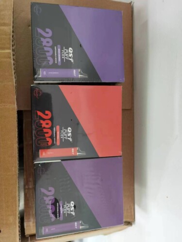 Trois boîtes scellées d'appareils de vape jetables QST Puff Flex, chacune offrant 2800 bouffées, avec les boîtes empilées dans un contenant en carton brun.