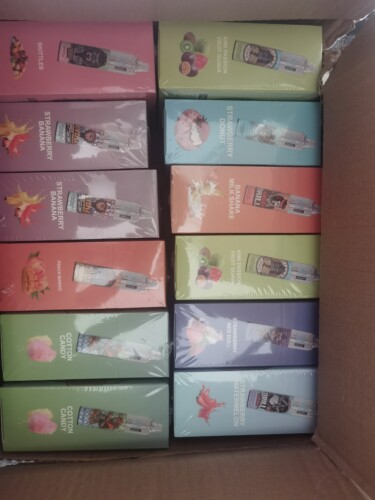 Une boîte contenant une variété de boîtes de jus de vape, chacune étiquetée avec différentes saveurs telles que Skittles, Kiwi Passion Fruit Guava, Strawberry Donut, Cotton Candy et Peach Mango, chacune accompagnée d'images de saveurs correspondantes.